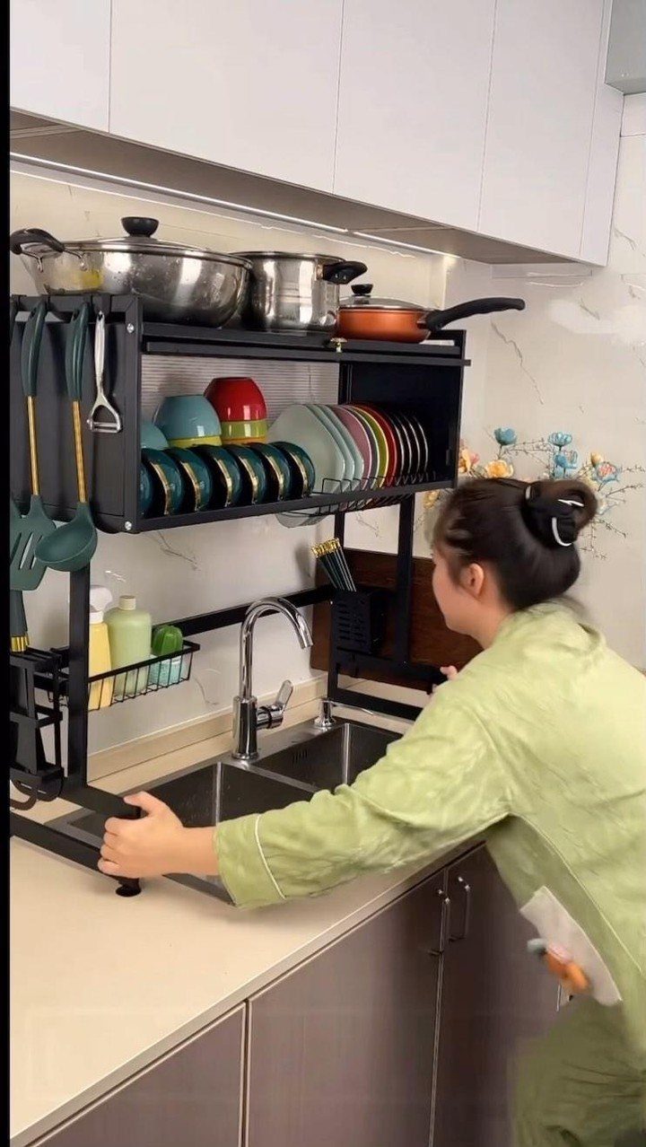 A person adjusting a black metal dish rack containing stacked colorful plates and bowls over a sink.