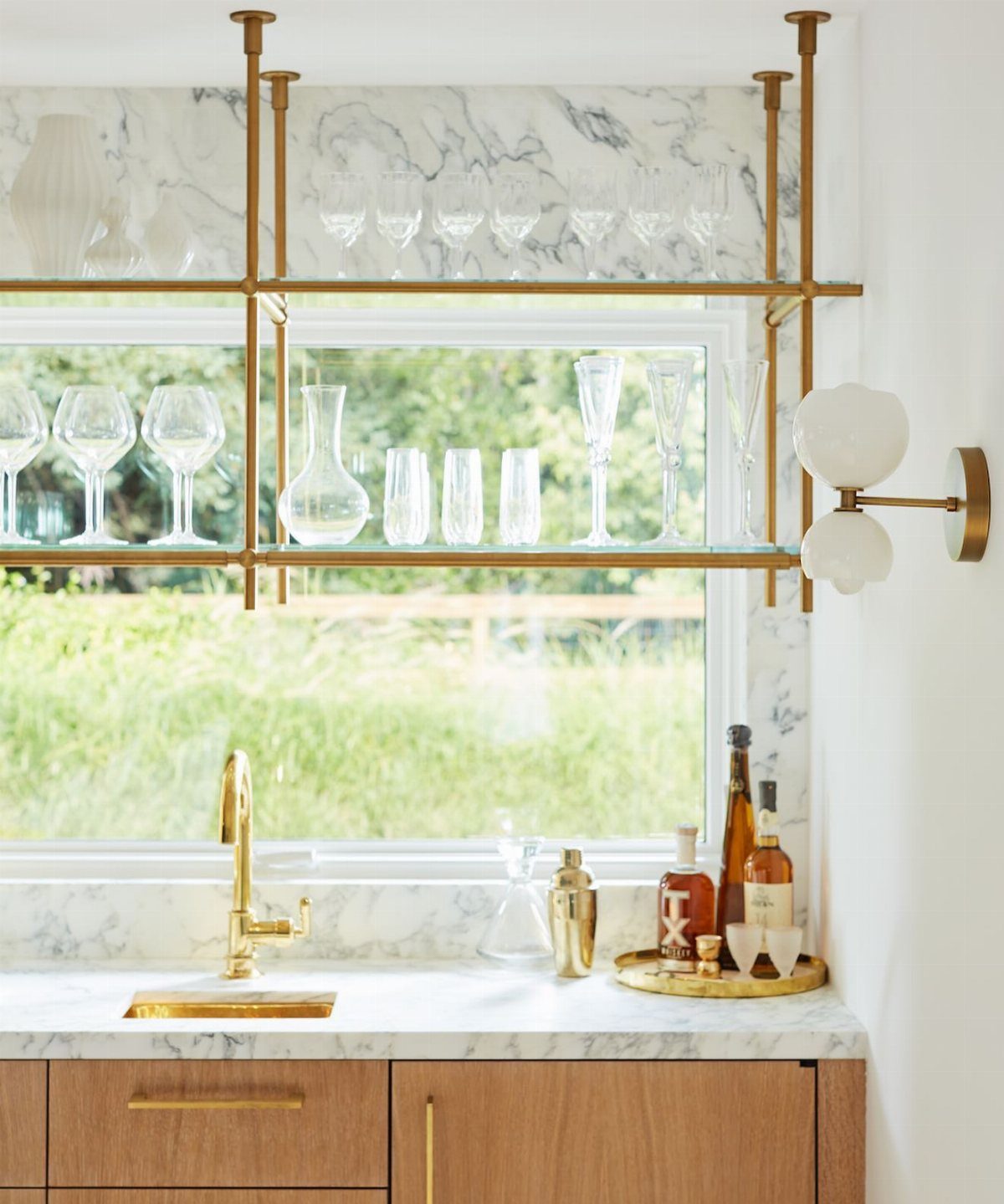 Gold metal shelving with glass shelves holding various clear glassware above a marble kitchen countertop.