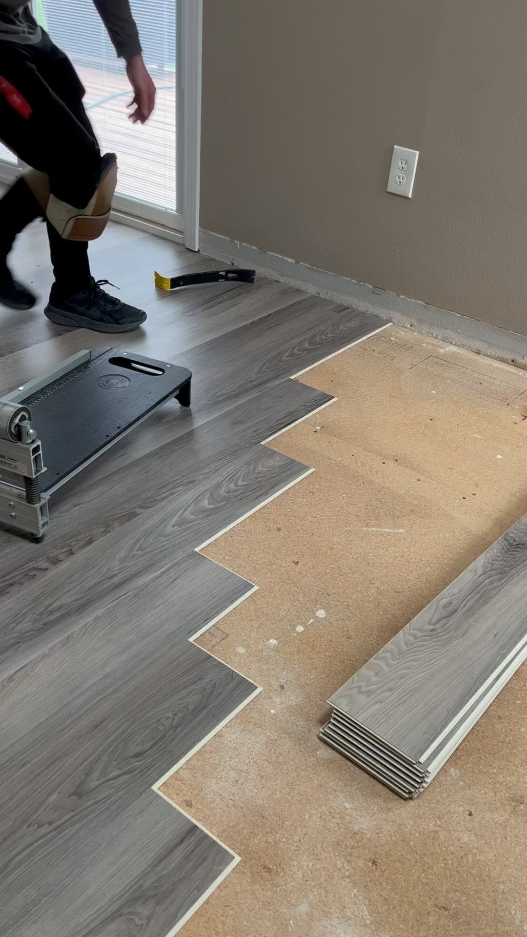 Person installing grey wood-textured laminate flooring planks over a light brown subfloor in a room.