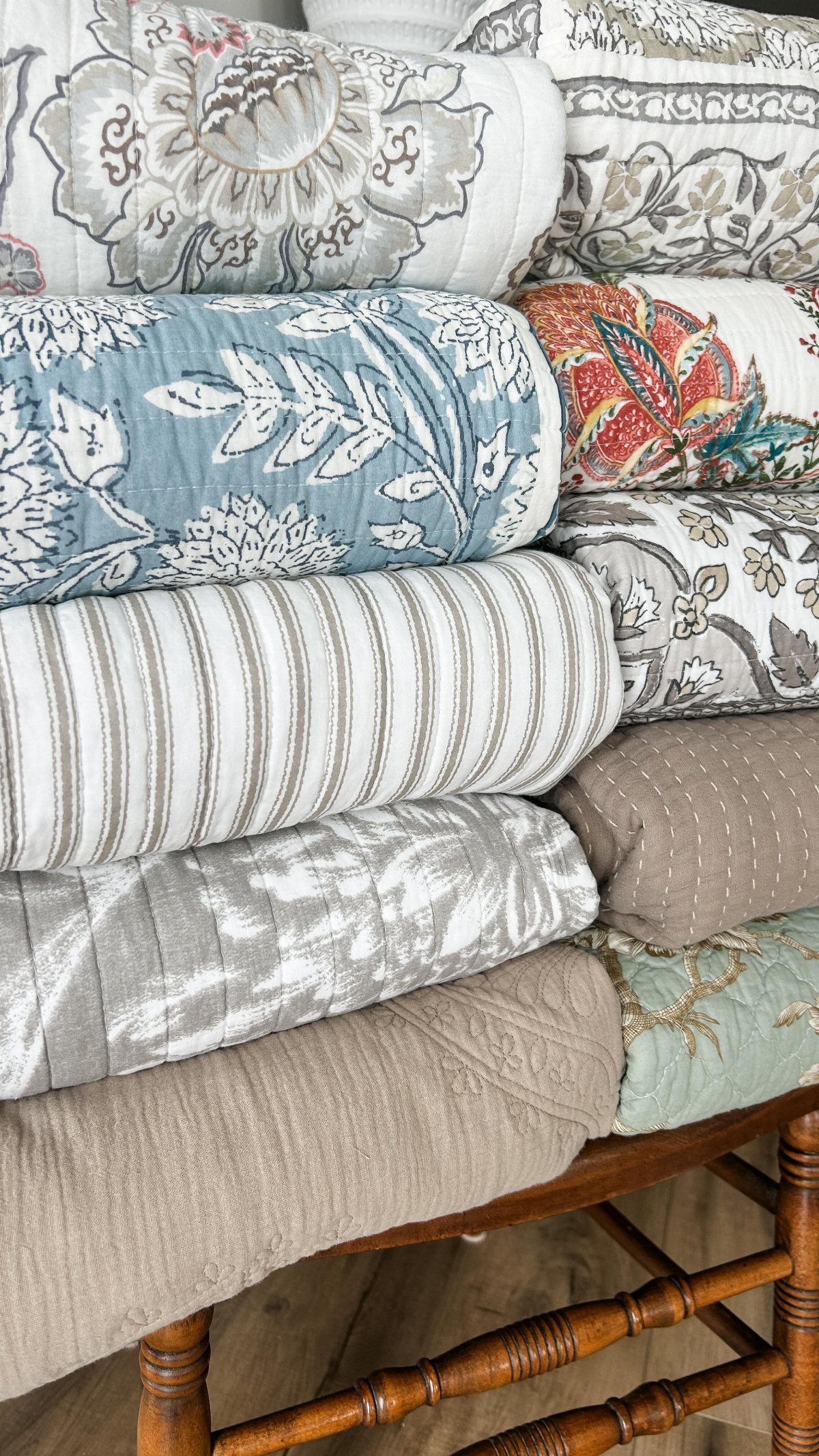 A stack of various patterned quilts and folded blankets in neutral and blue tones.