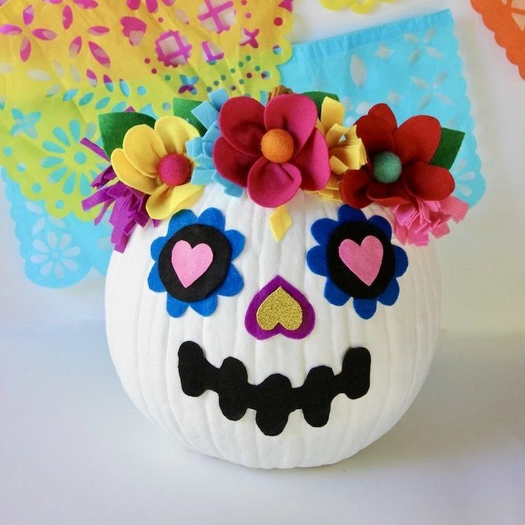 White sugar skull pumpkin decorated with colorful felt flowers and heart-shaped eye designs.