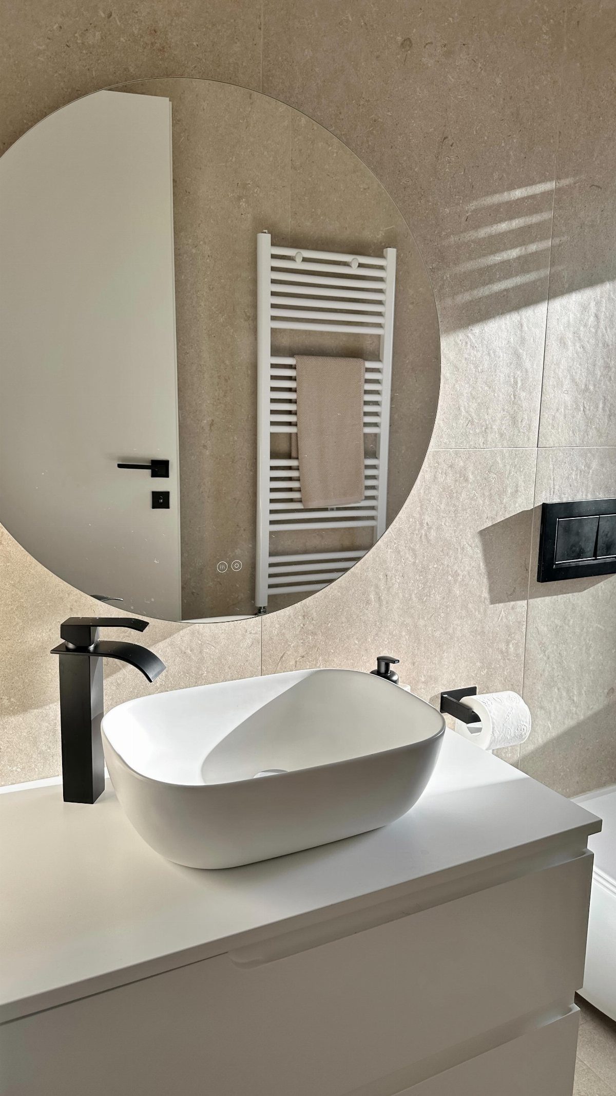 White vessel sink and circular mirror set against beige stone tiles in a sunlit bathroom.