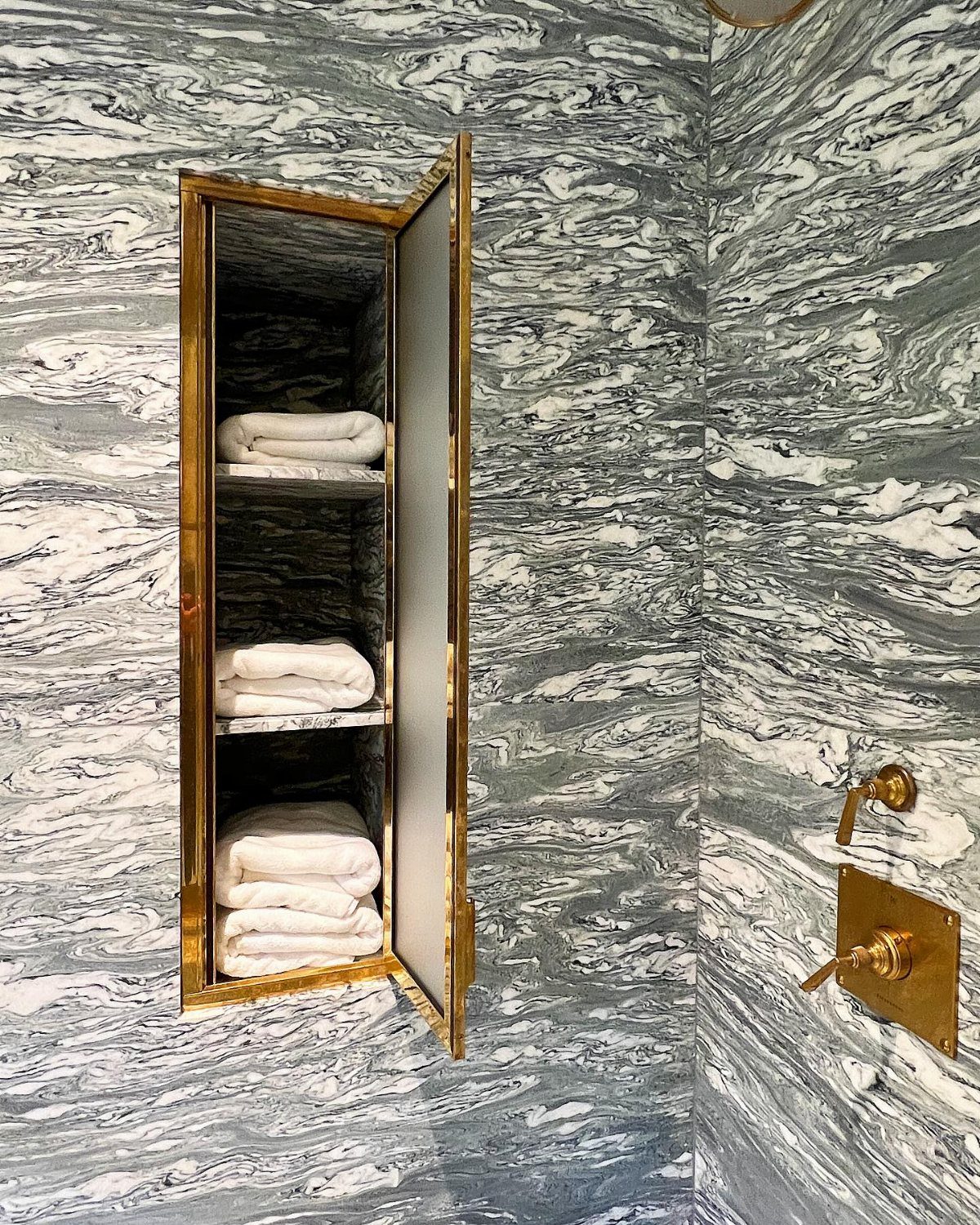 Folded white towels stacked inside a gold recessed wall cabinet against green marble tiles.