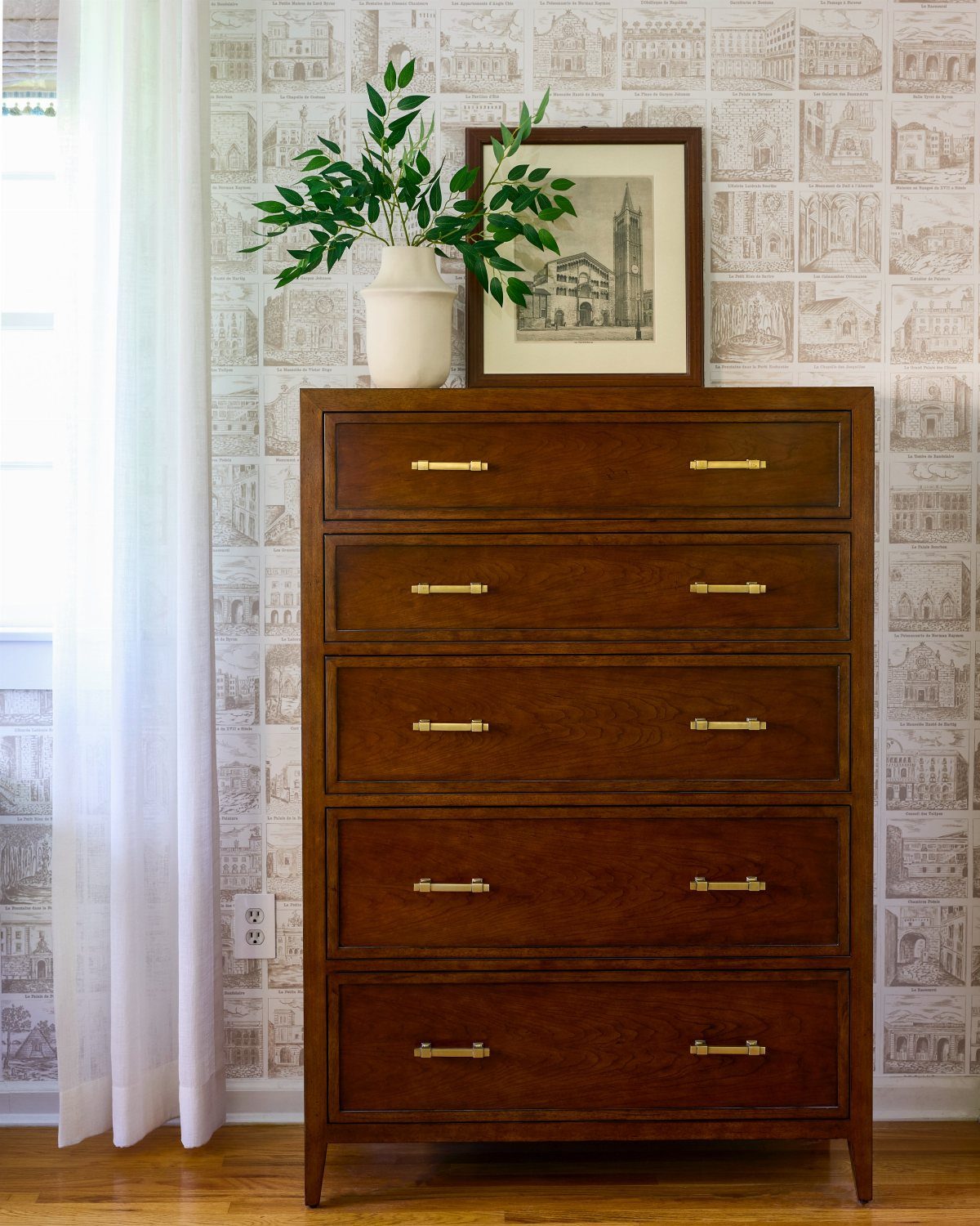 A dark wood dresser with gold handles sits against patterned wallpaper with greenery and art.
