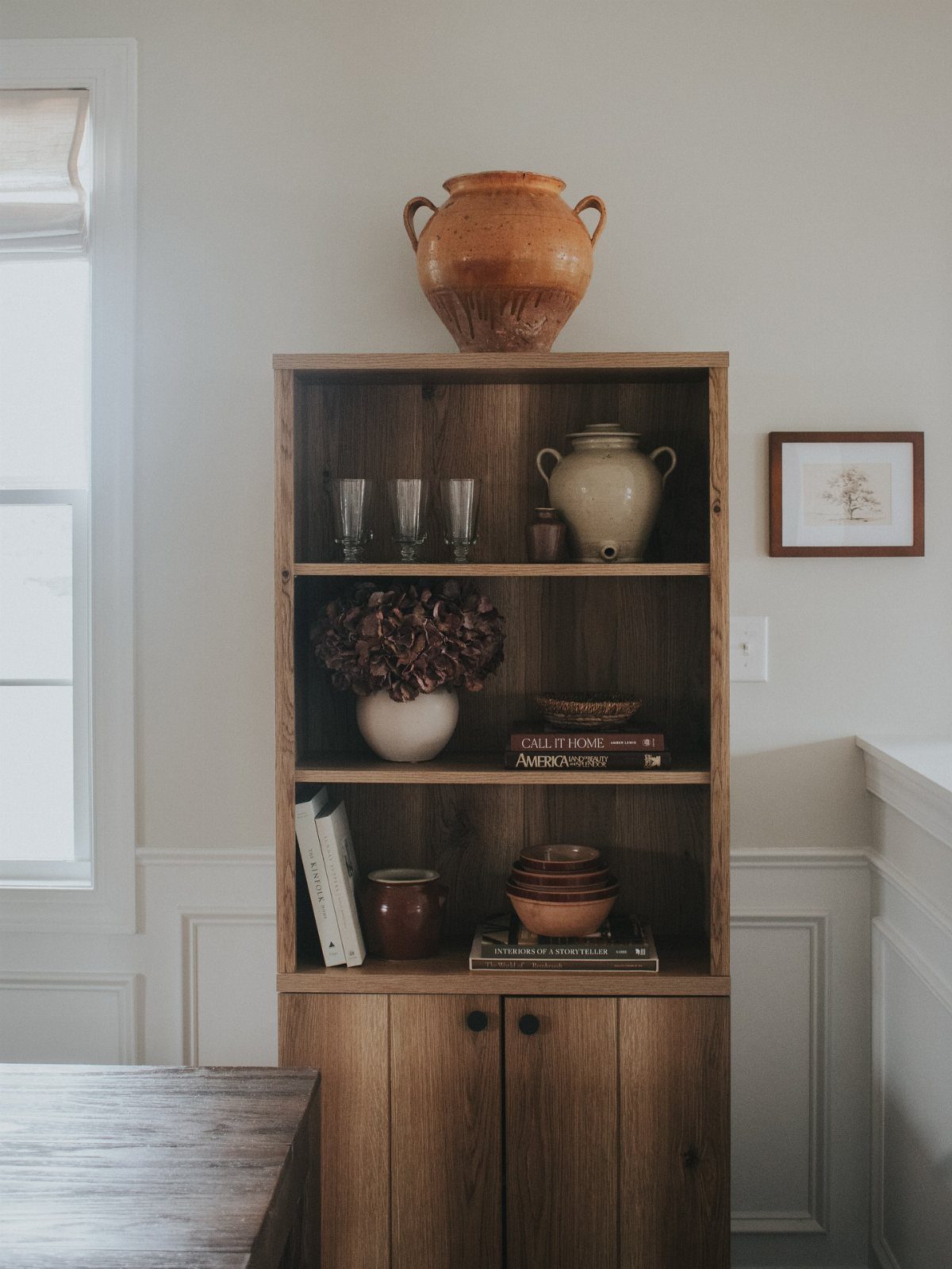 Wooden bookshelf displaying curated ceramic vases, bowls, and books against a neutral wall.