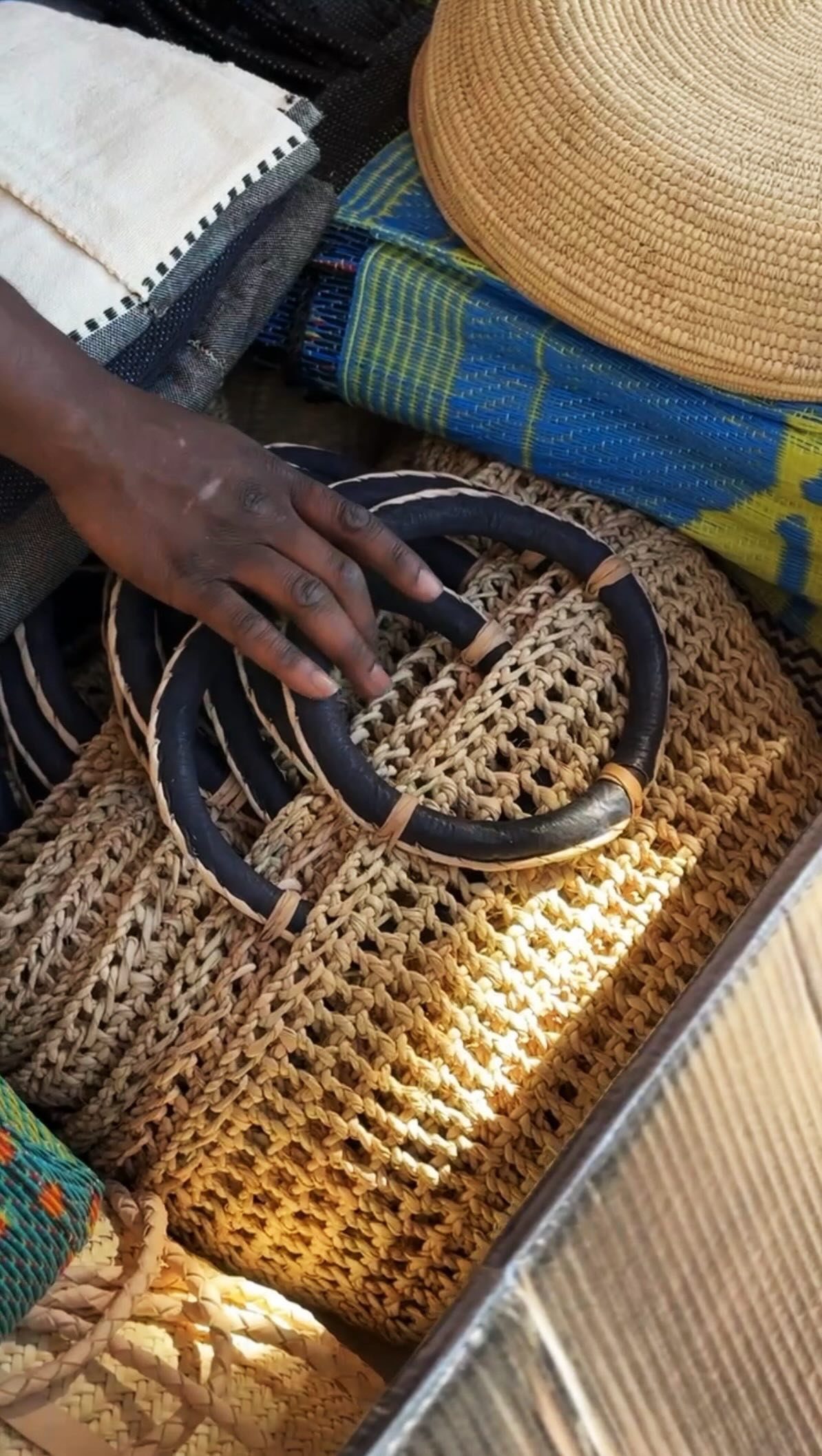 A hand touching a large woven straw basket with black handles filled with textiles.