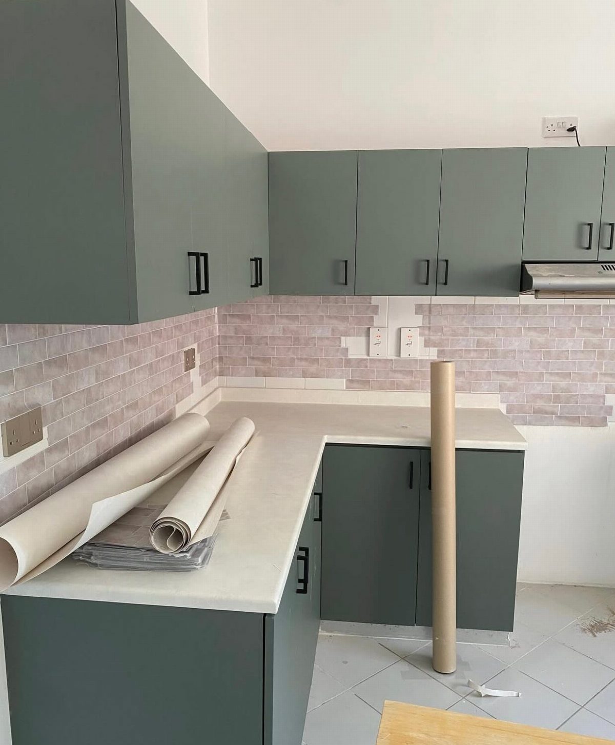 Green kitchen cabinets with a brick-patterned backsplash and rolls of wallpaper on the counter.