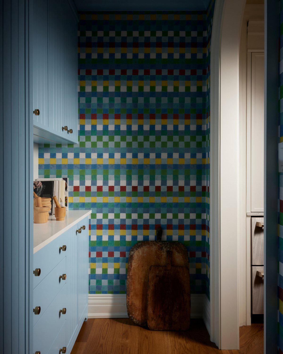 Colorful geometric checkered wallpaper in a small pantry with blue cabinets and wooden flooring.