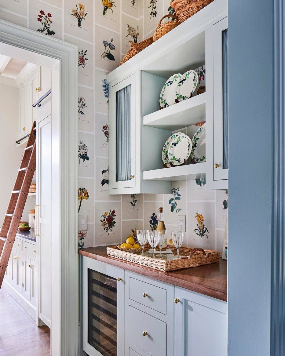 Floral patterned wallpaper behind light blue kitchen cabinetry and open shelving with decorative plates.