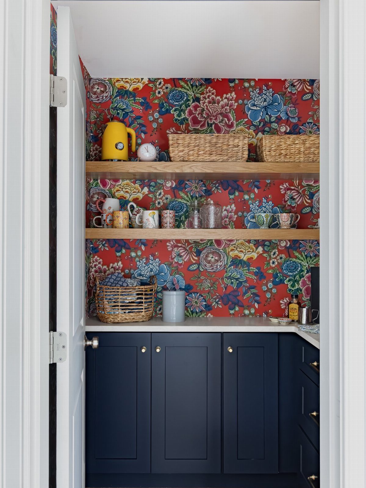 Red floral wallpaper decorating the walls of a pantry with wooden shelves and navy blue cabinets.