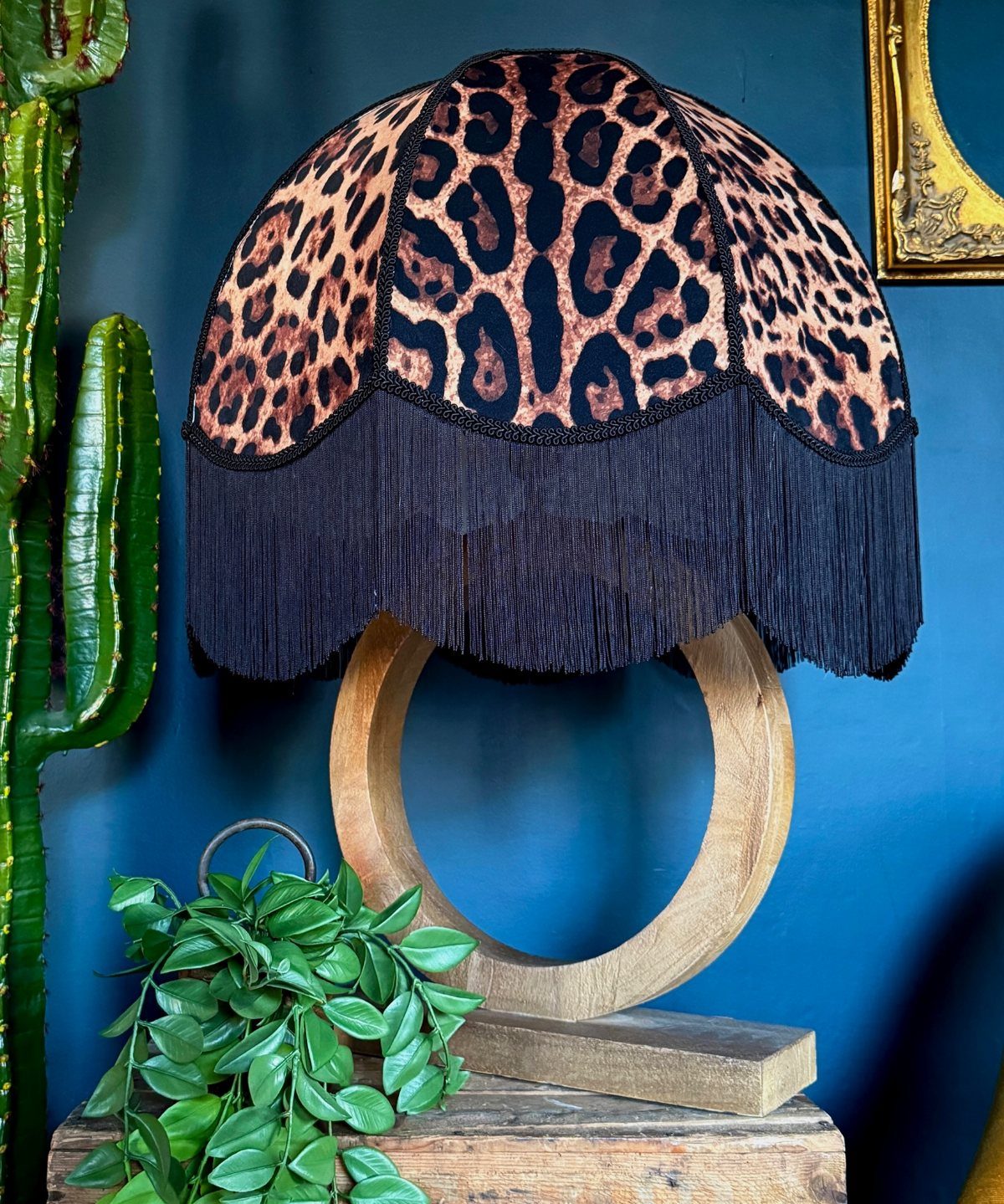 Leopard print lampshade with black fringe on a wooden circular base against a blue wall.