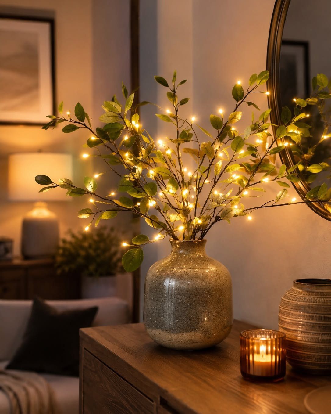 Leafy branches with warm fairy lights in a ceramic vase on a wooden table.