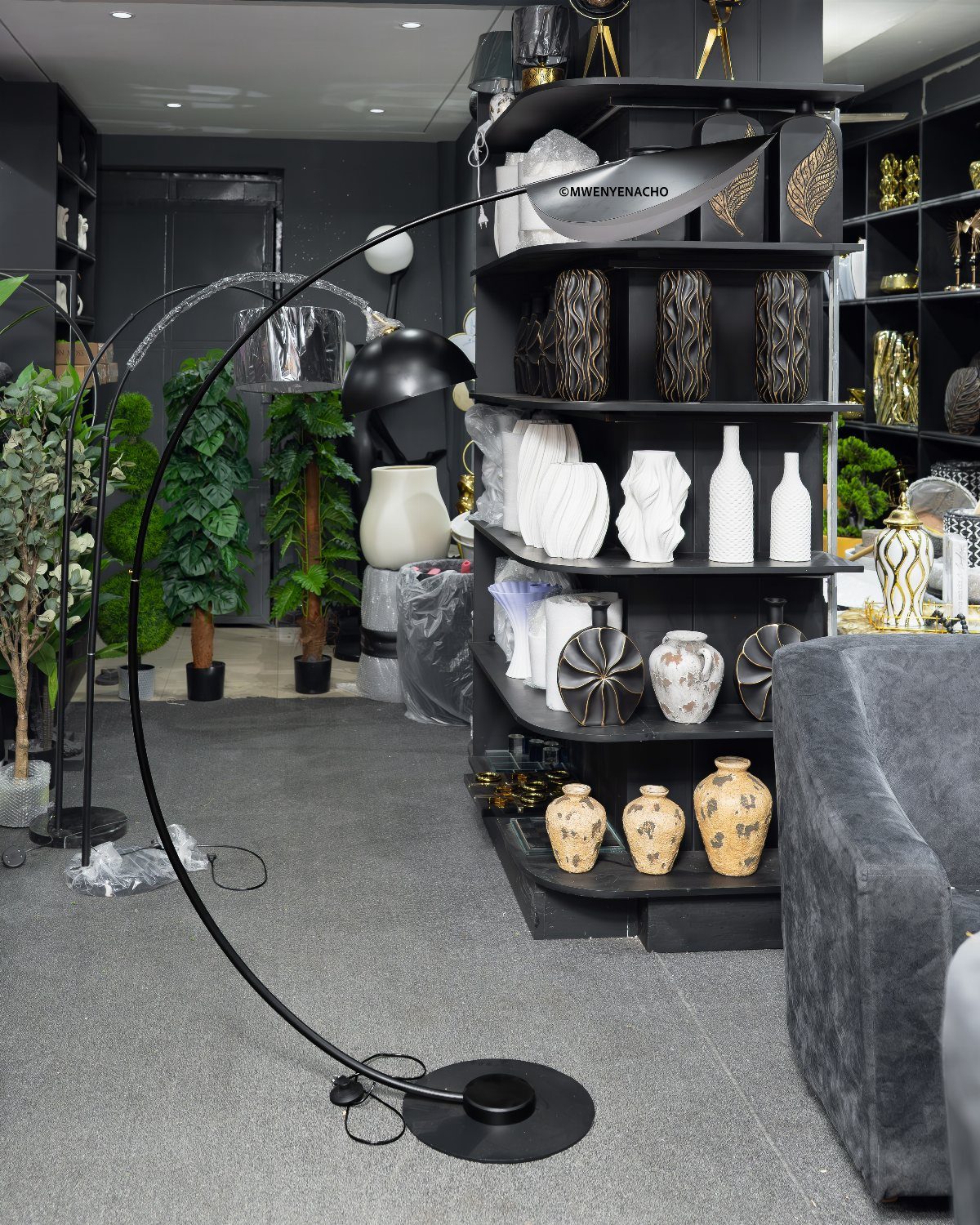 Black arched floor lamp standing in a modern showroom filled with decorative vases and plants.