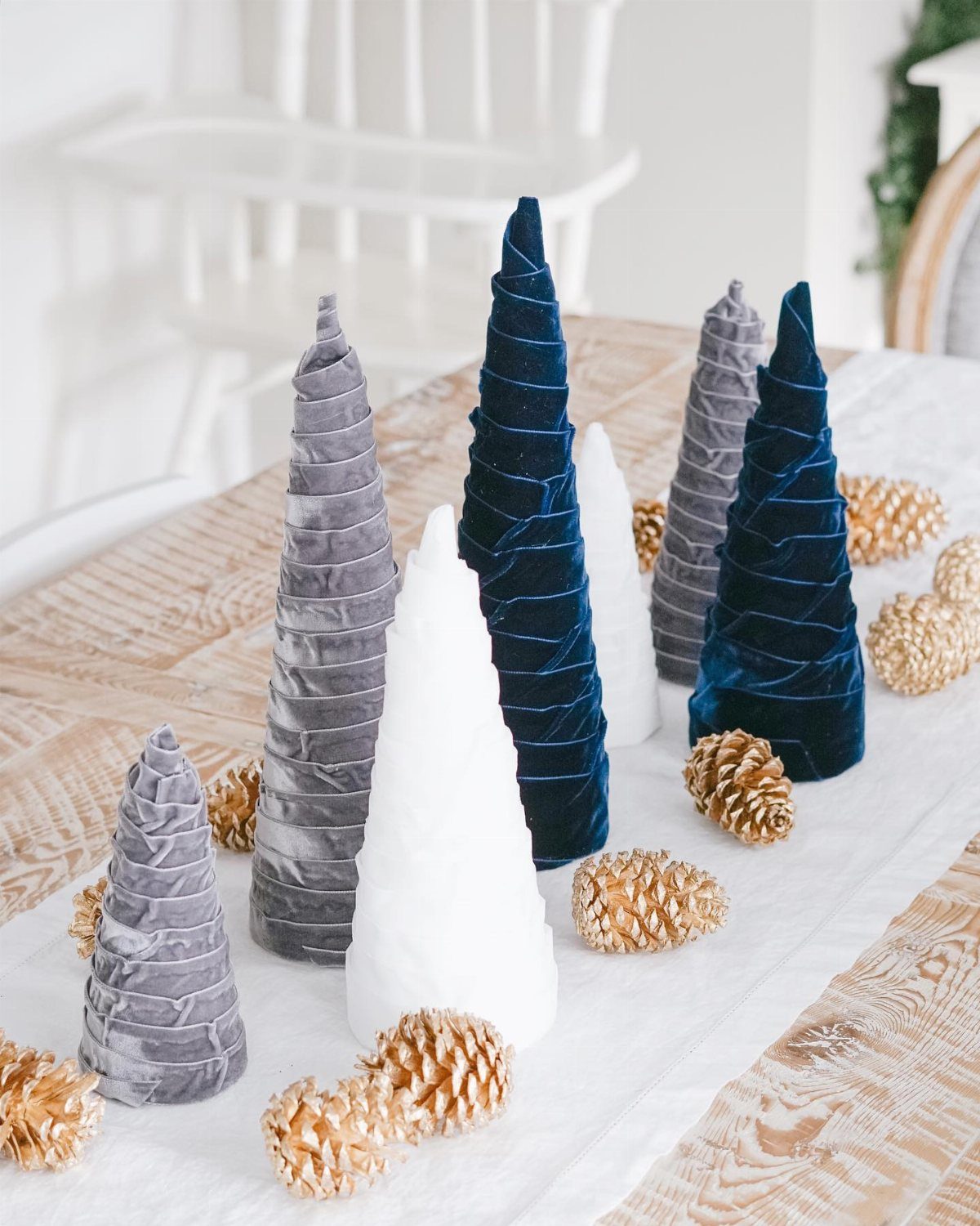 Small velvet conical trees in grey, white, and navy blue arranged with gold pinecones on a table.