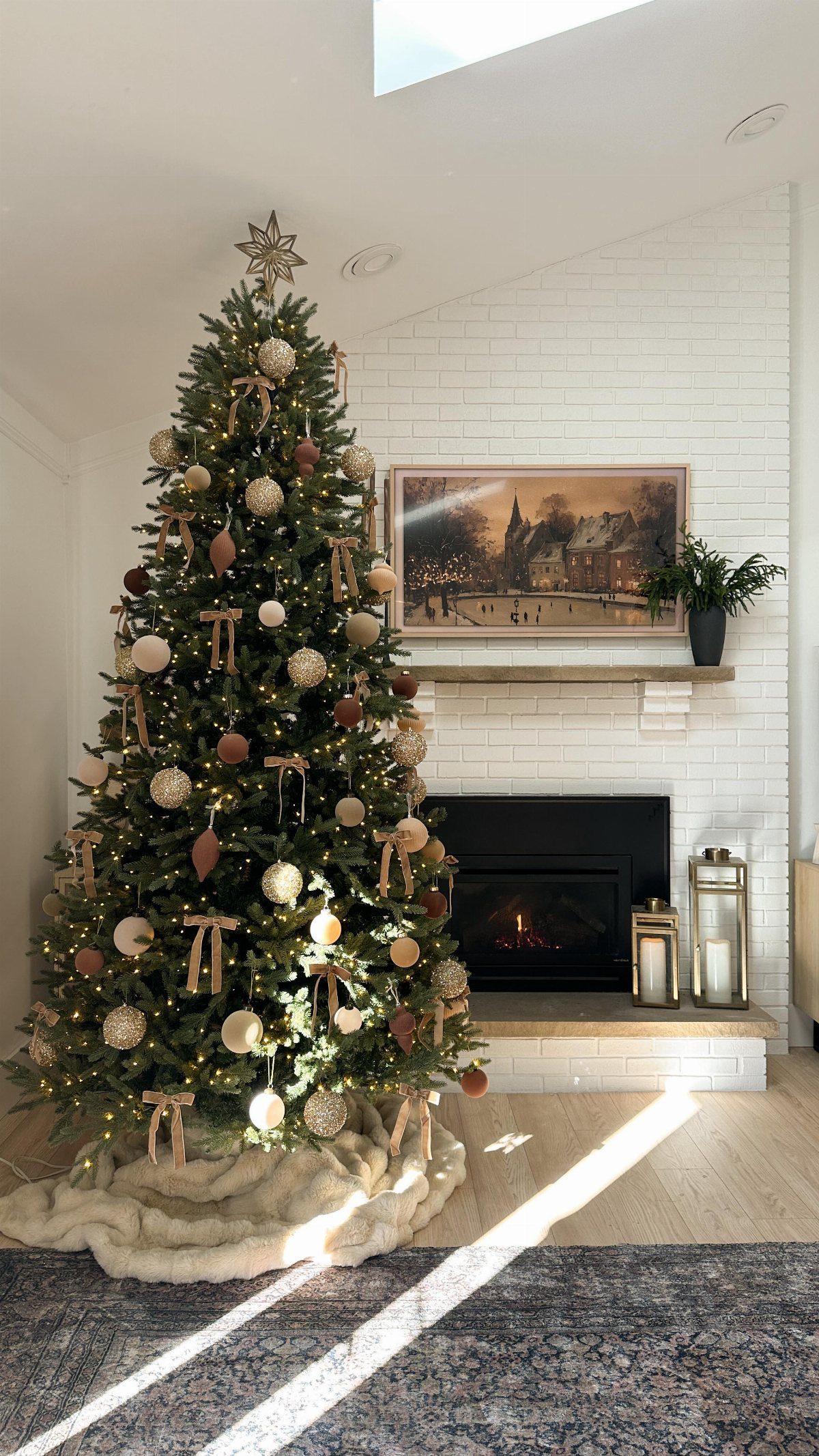 Decorated Christmas tree with neutral ornaments and fairy lights standing near a white brick fireplace.