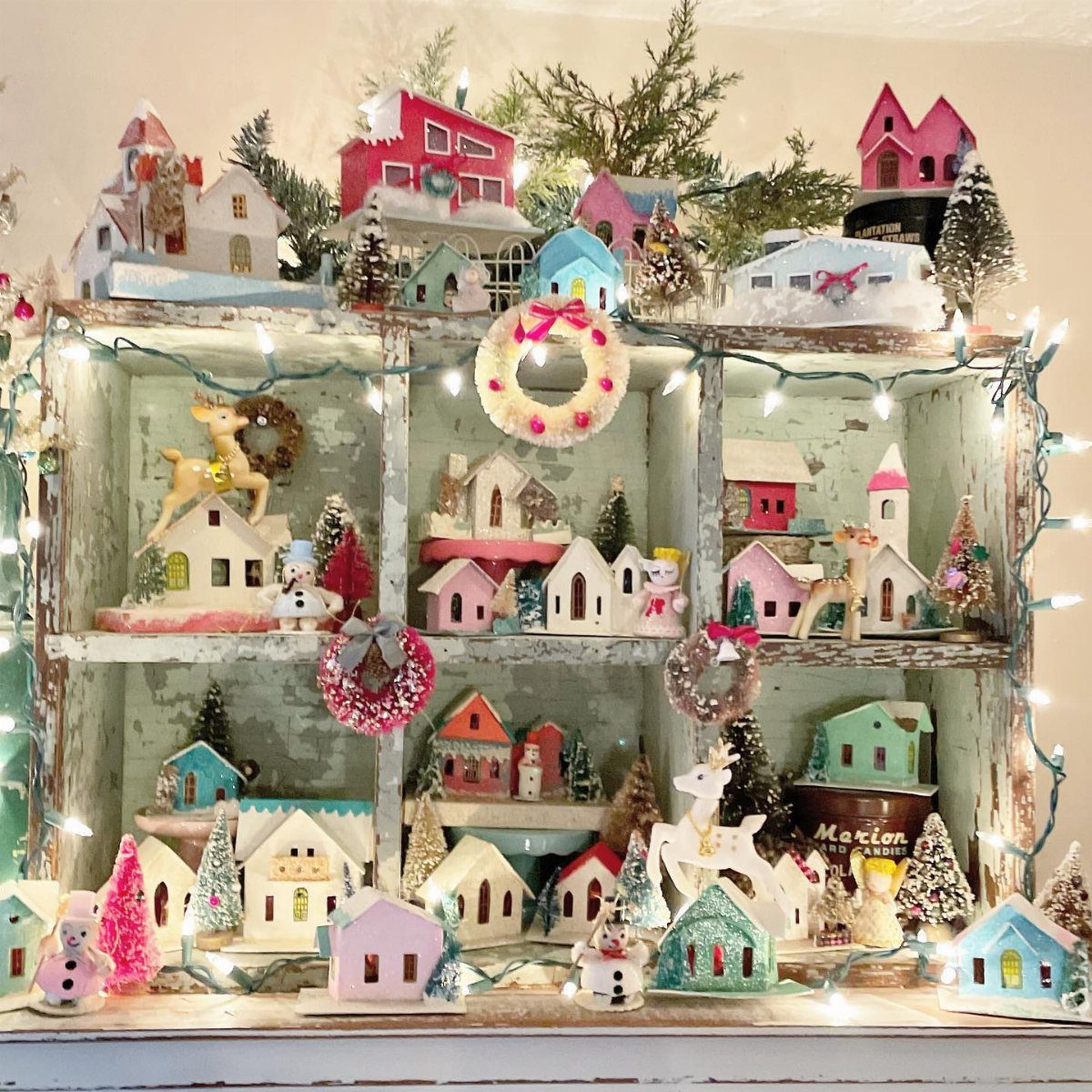 Miniature colorful houses and festive decorations arranged on a light blue wooden shelf with fairy lights.