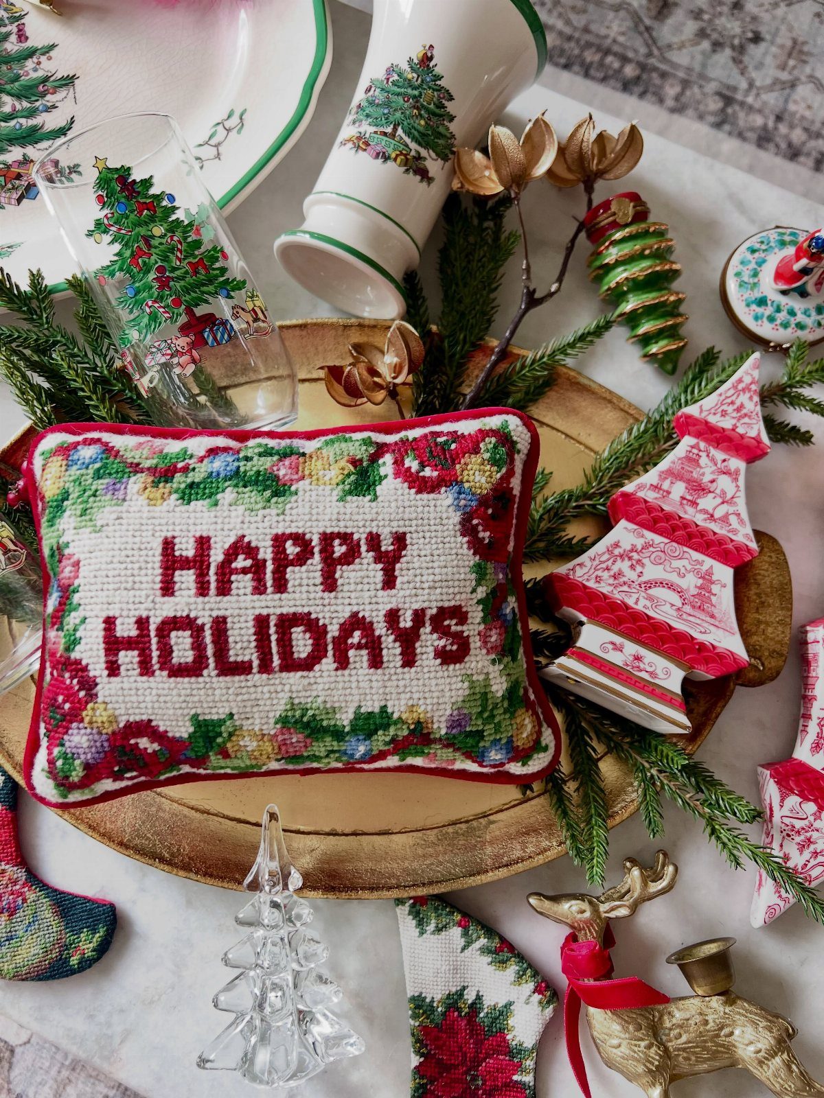 Cross-stitch holiday pillow and vintage-style ceramic trees arranged on a gold tray with greenery.