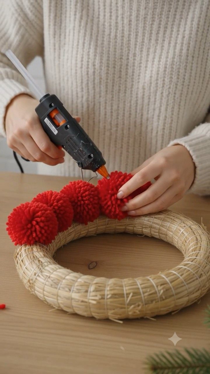 Person using a hot glue gun to attach red pom-poms to a straw wreath.