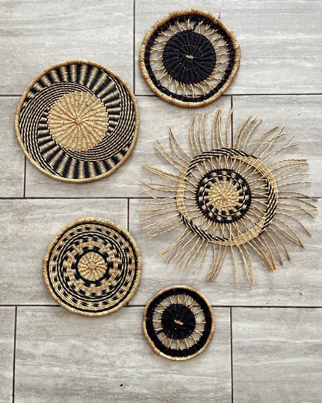 Five woven circular baskets with black and natural patterns arranged on a grey tiled wall.