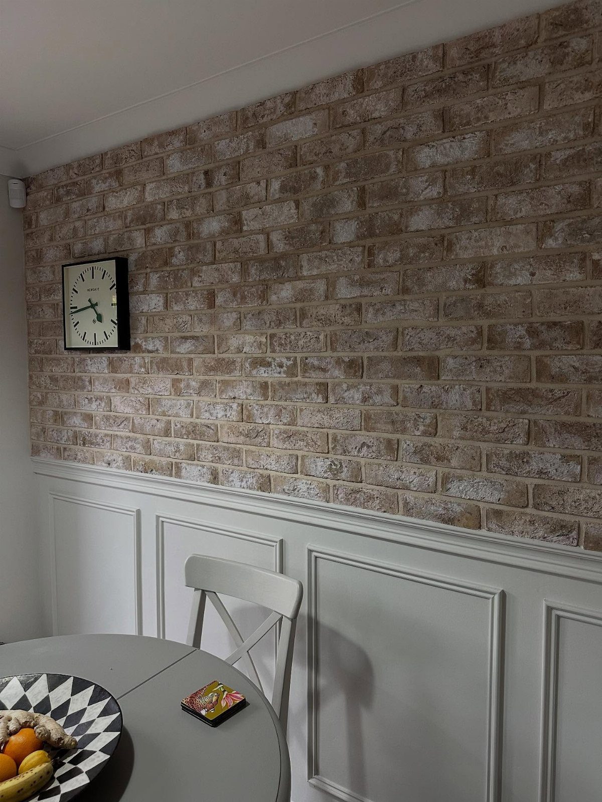 Faux brick patterned wall above white wainscoting with a small black clock hanging on it.
