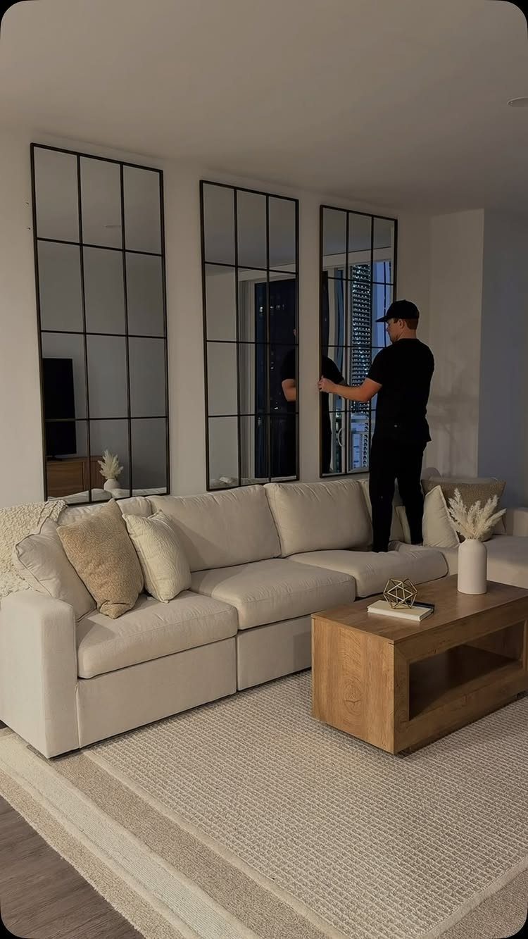 A man installs three large black-framed paneled mirrors on a white living room wall.