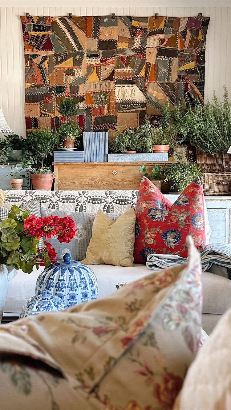 A large multicolored patchwork tapestry hanging on a white paneled wall above indoor plants.