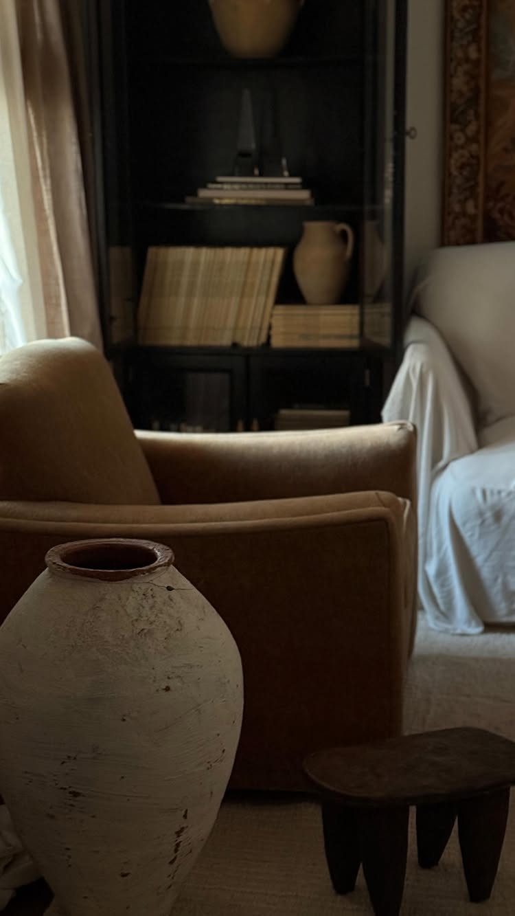 Large white ceramic vase in front of a tan armchair and dark bookshelf.