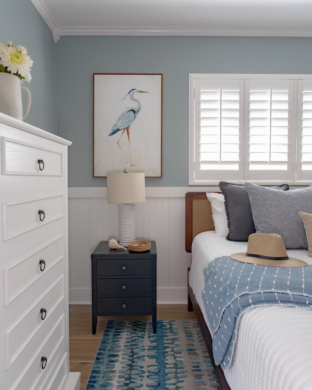 A coastal bedroom with blue walls, white shutters, a bed with blue textiles, and heron artwork.