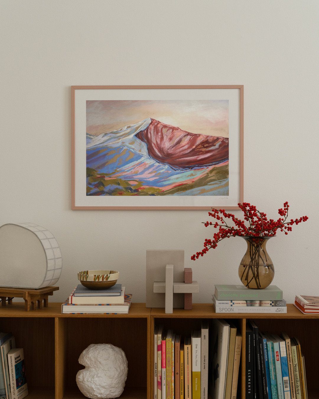A framed mountain landscape painting hangs above a wooden bookshelf filled with books and decor.