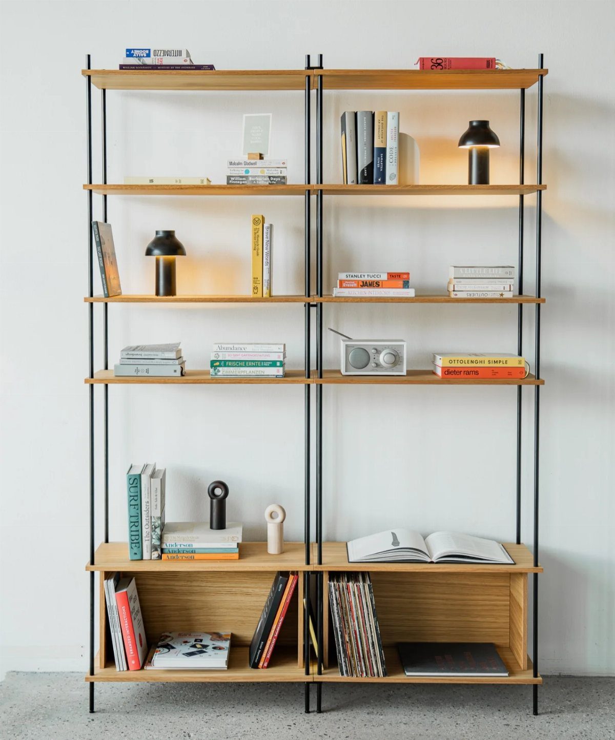A minimalist wooden bookshelf with black metal frames holding books and small lamps.