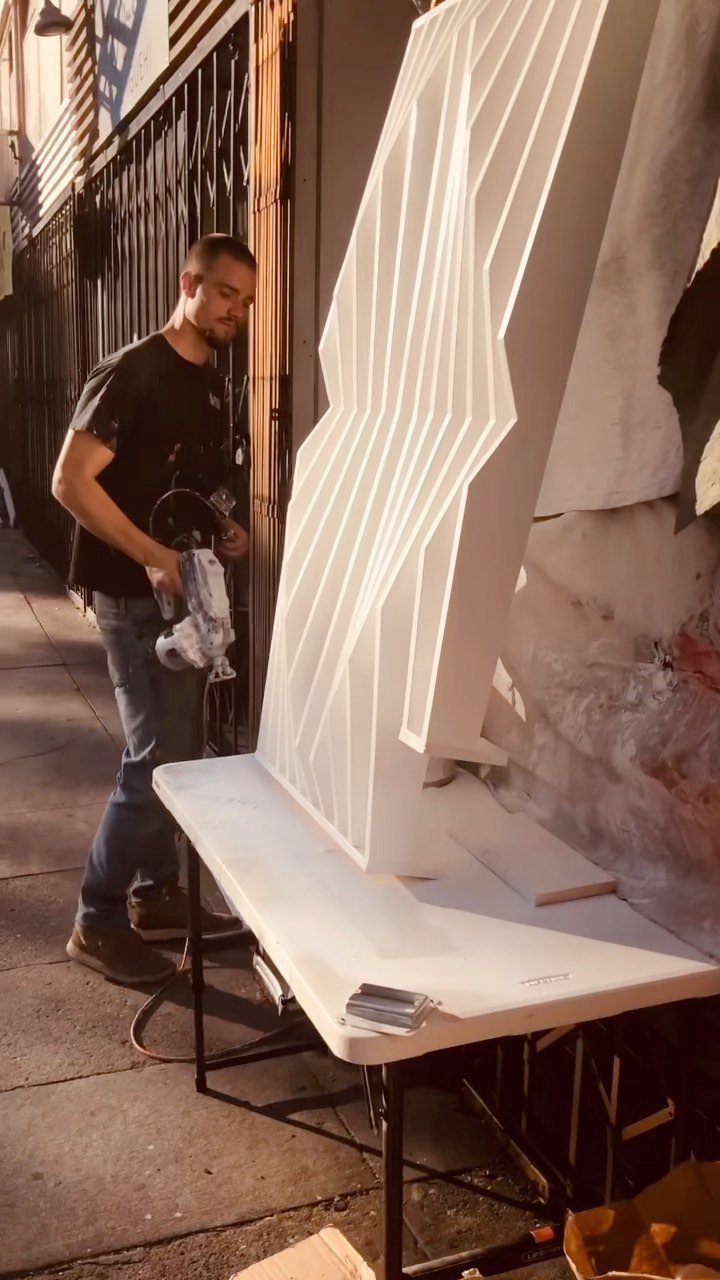 A man spraying white paint onto a large, angular, three-dimensional geometric decorative panel.