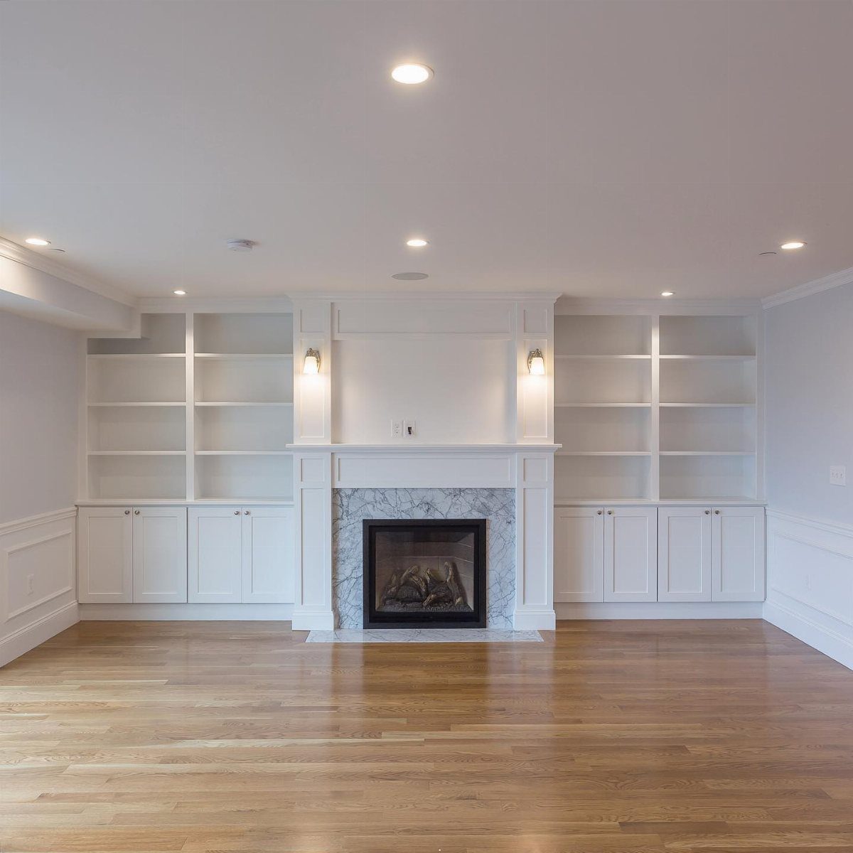 Empty living room featuring white built-in shelving and a marble fireplace with wood floors.