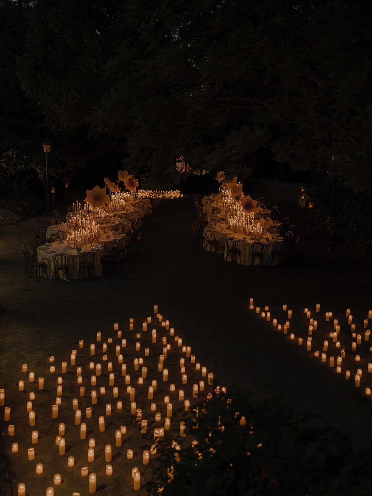 Rows of glowing candles on tables and ground illuminating an outdoor evening dining setting.