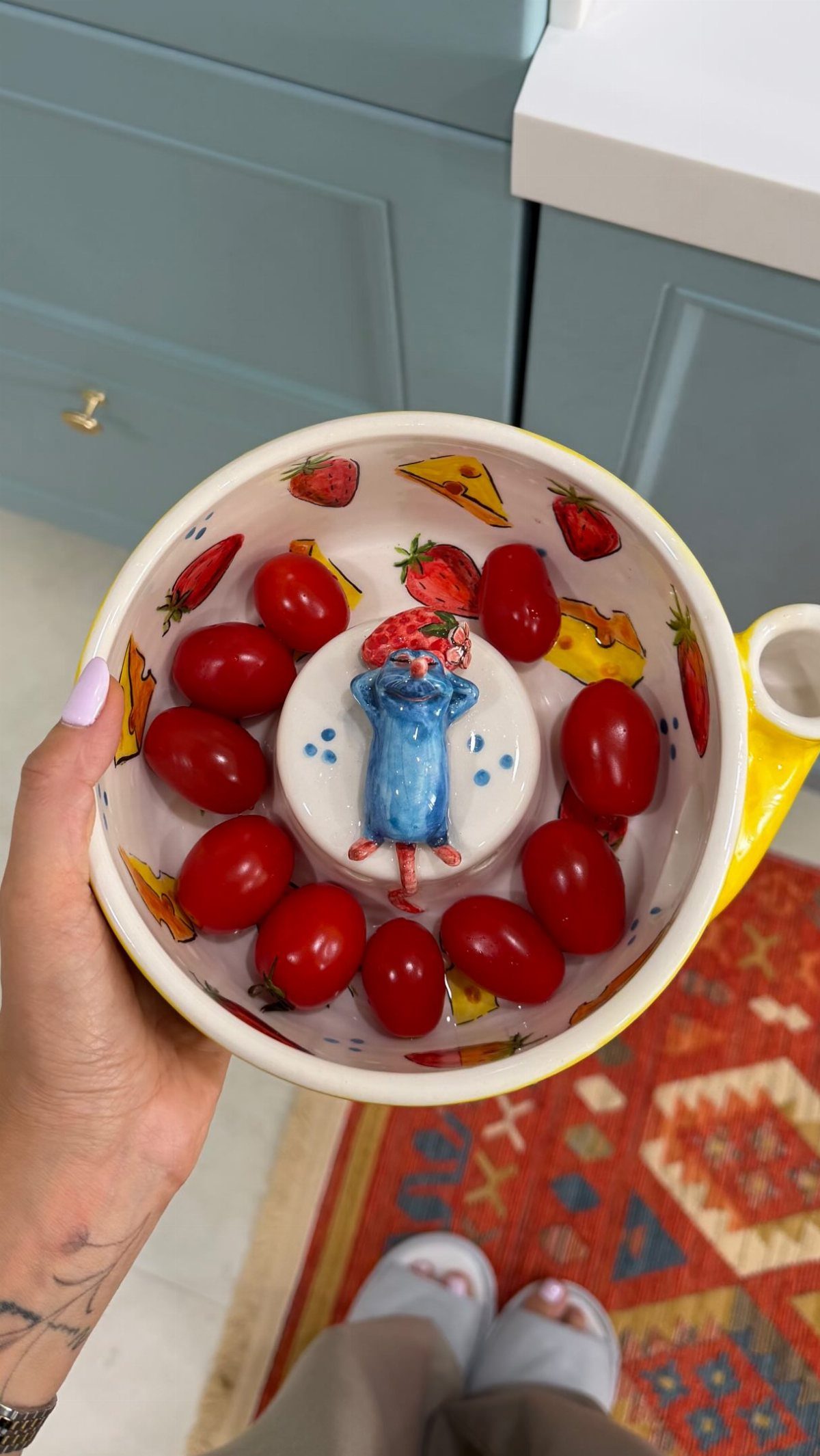 A hand holding a decorated ceramic bowl filled with small red tomatoes and a blue mouse figurine.