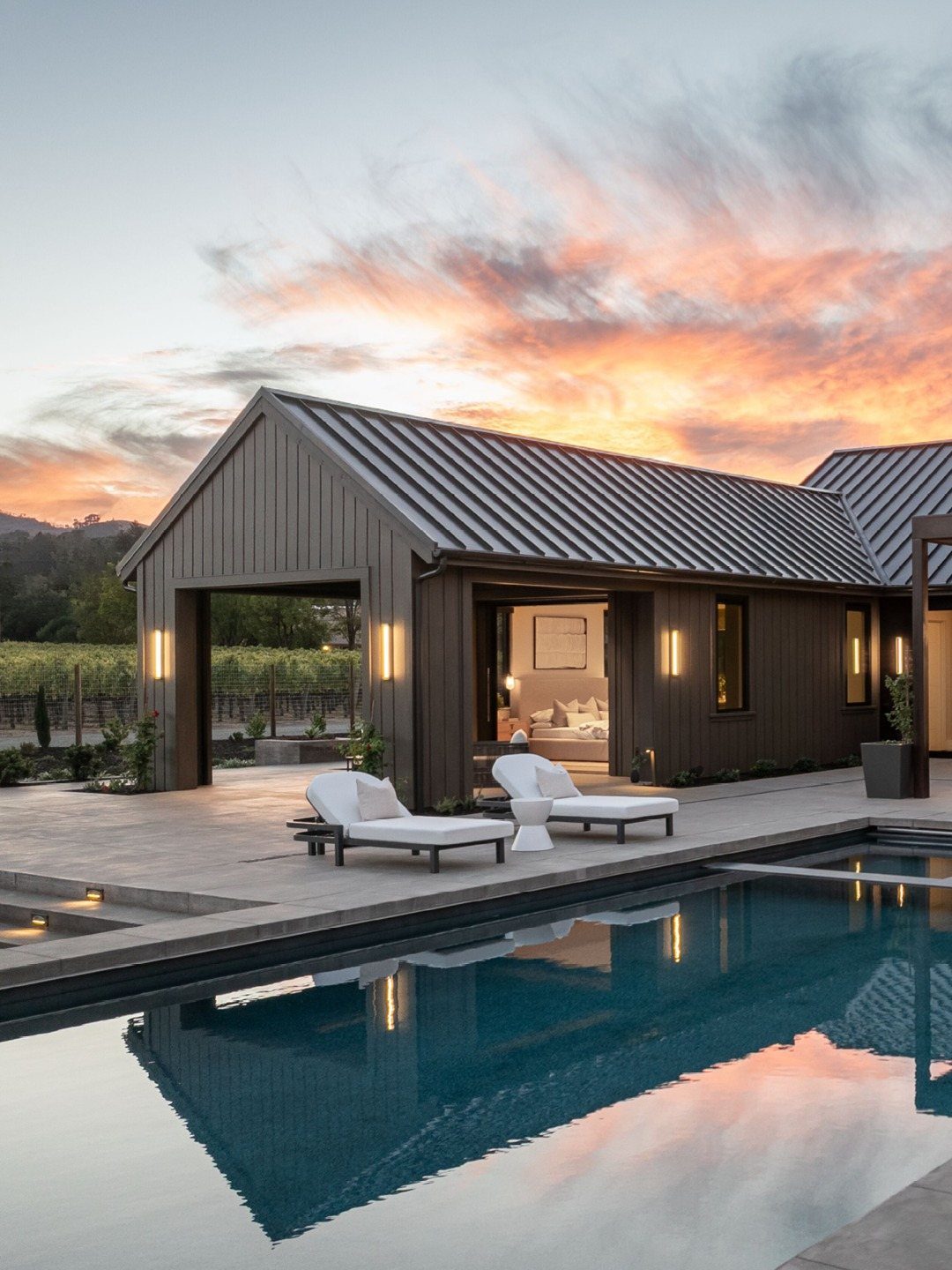 Modern dark wood house with open living spaces, white lounge chairs, and a swimming pool at sunset.