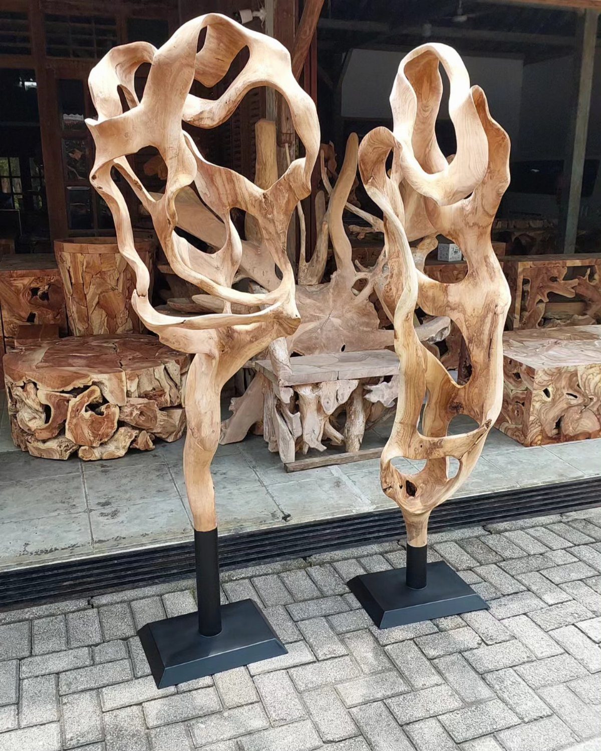 Two large, organic wooden sculptures with hollowed patterns standing on black bases outdoors.