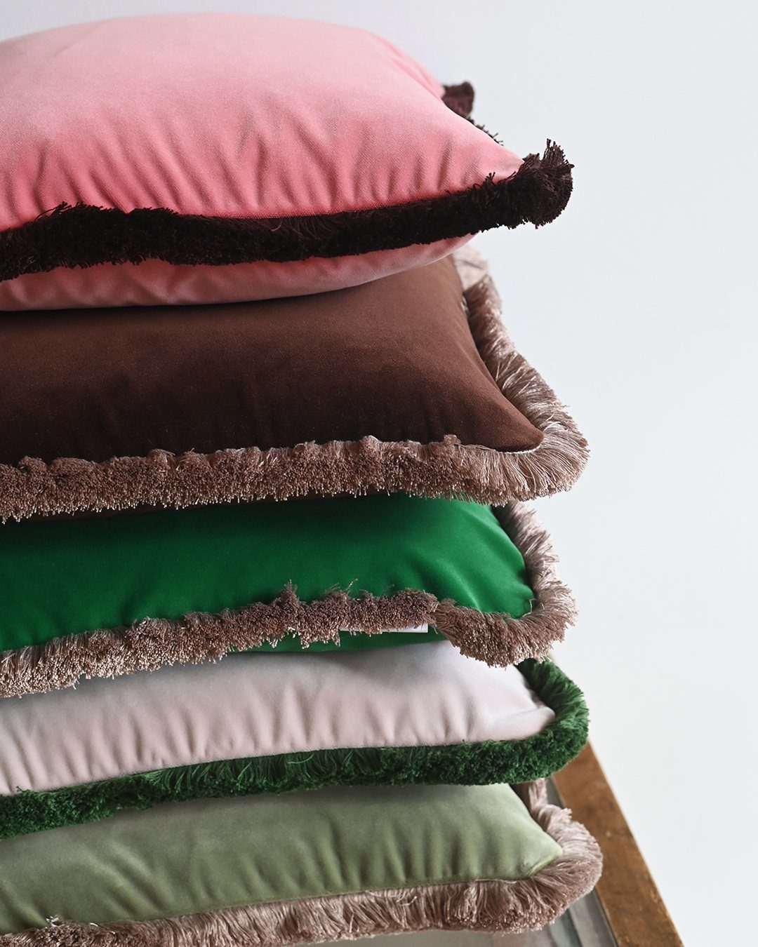 A stack of colorful velvet cushions with decorative fringed edges in pink, green, and brown.