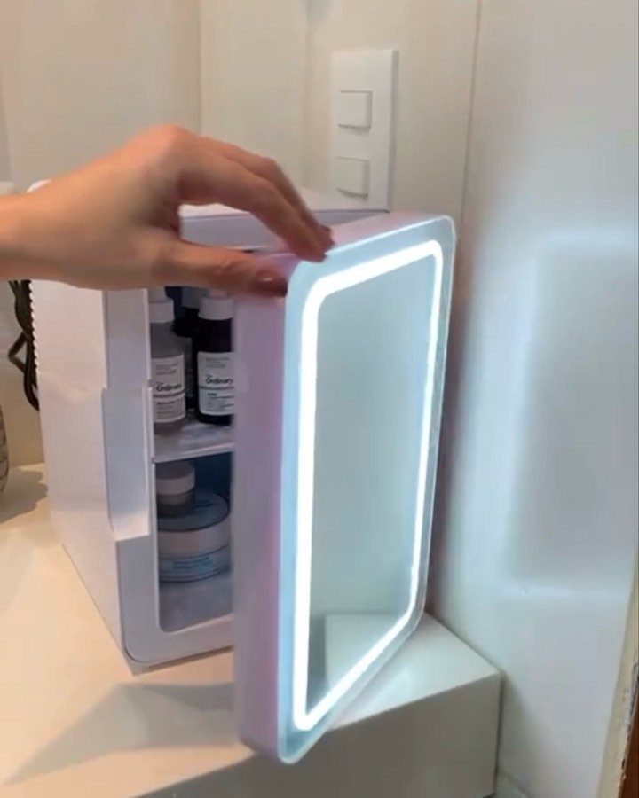 A hand opens a small glowing skincare mini fridge containing various beauty products.