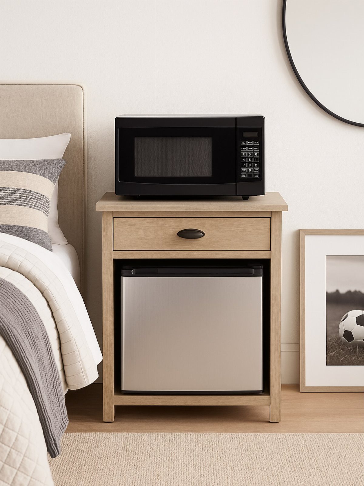 A light wood nightstand holding a black microwave with a silver mini fridge underneath.