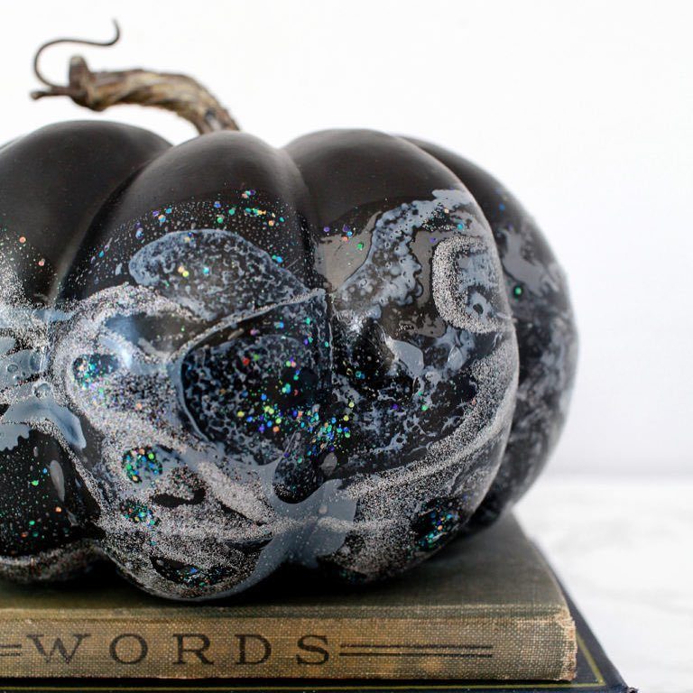 Black pumpkin decorated with swirling silver glitter and iridescent cosmic speckles sitting on a book.