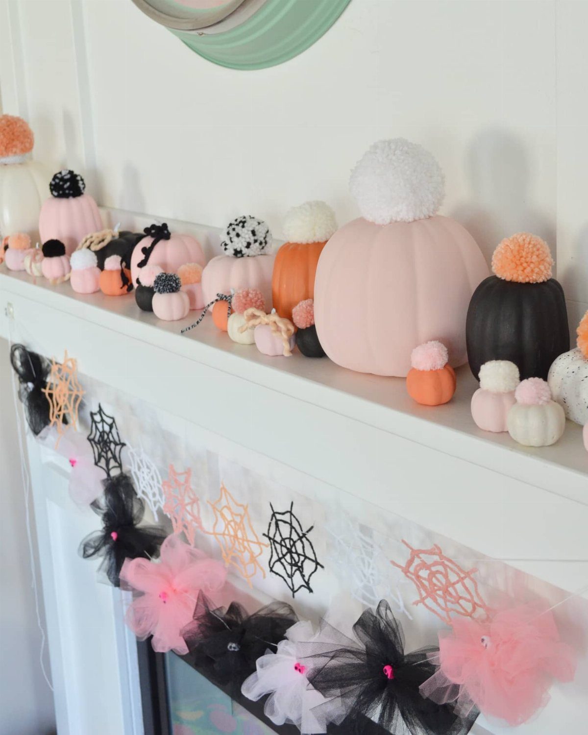 Various pastel painted mini pumpkins decorated with colorful pom poms sitting on a white mantelpiece.