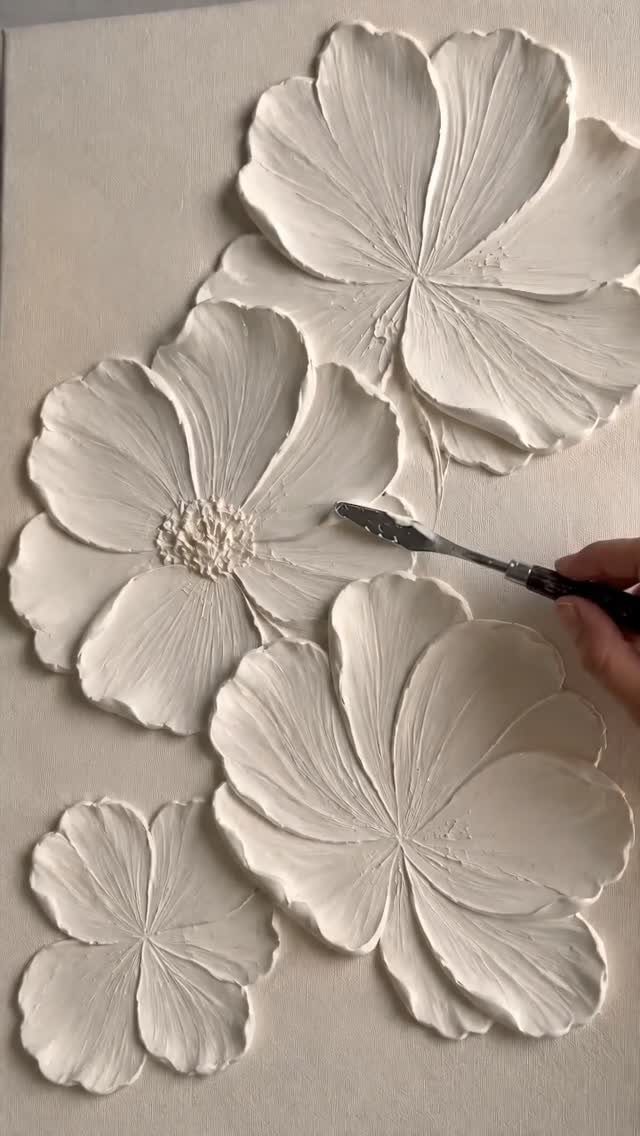 A hand uses a palette knife to sculpt thick white plaster flowers onto a canvas.