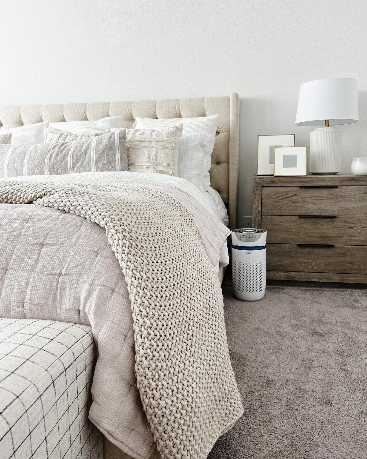 A minimalist bedroom with beige tufted headboard, neutral layered bedding, and a wooden nightstand.