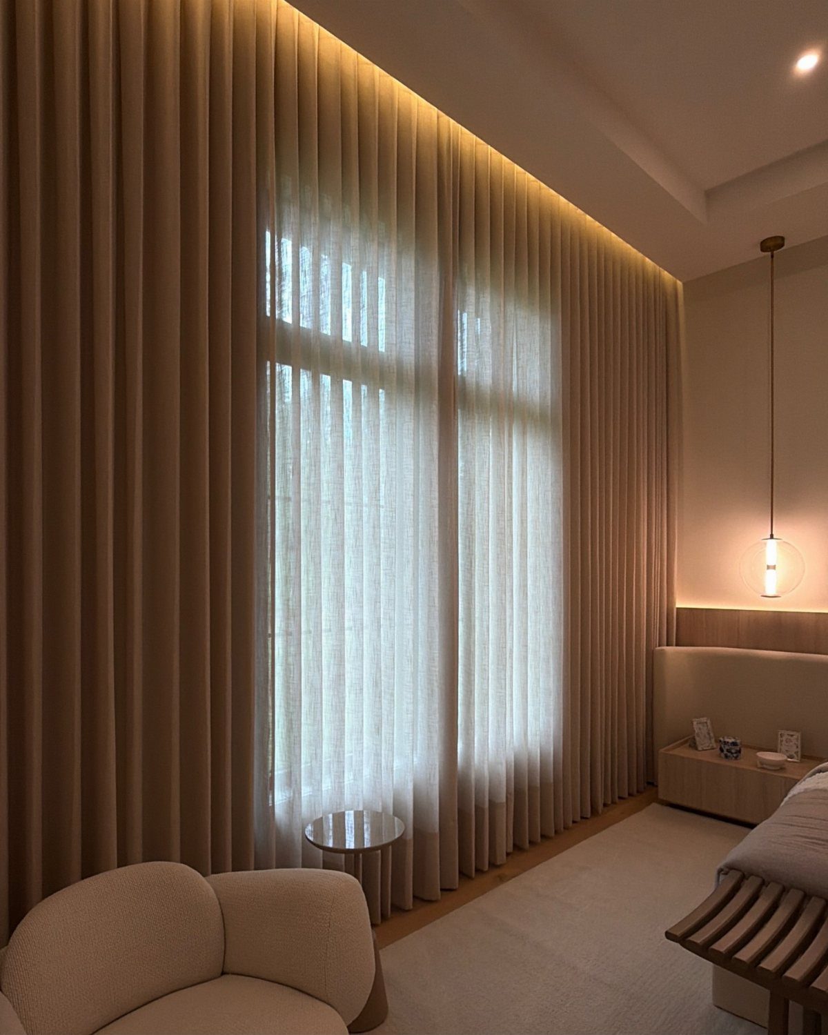 Beige floor-to-ceiling sheer curtains hanging in a bright, minimalist bedroom with warm lighting.