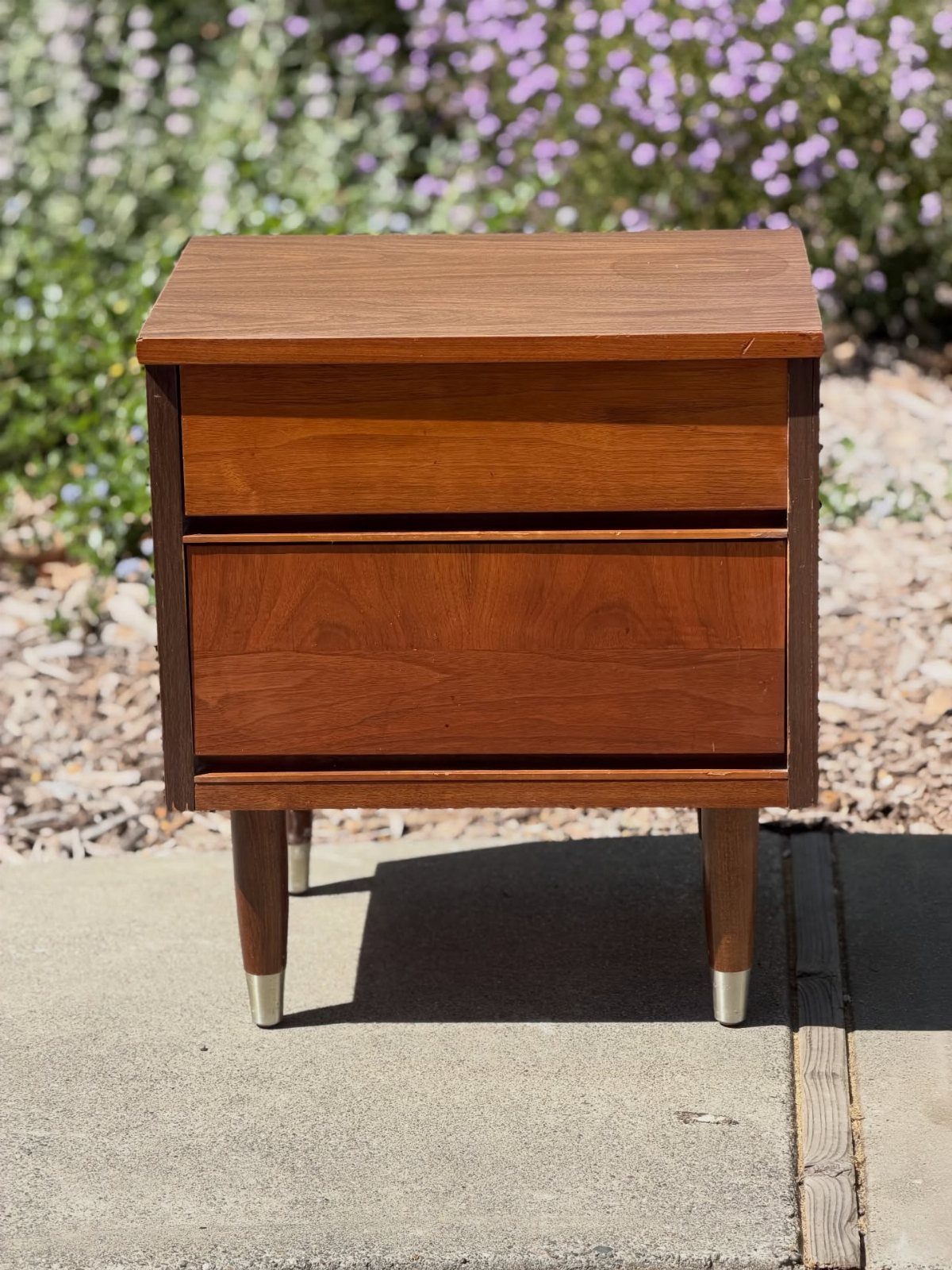 Mid-century modern wooden nightstand with two drawers and gold-tipped tapered legs outdoors.