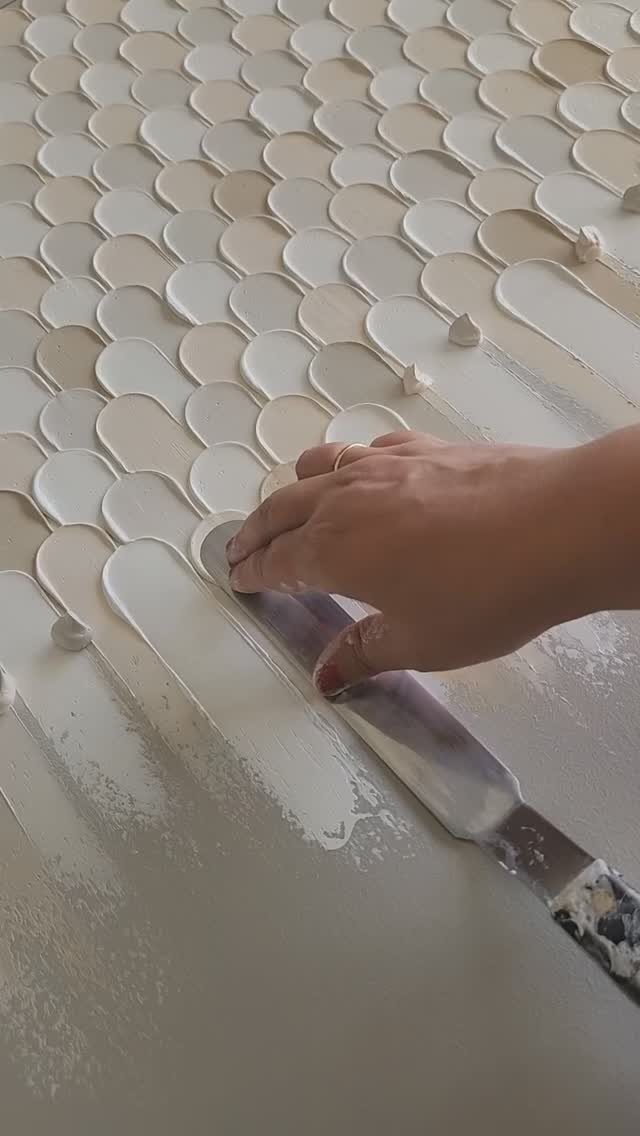 A hand uses a spatula to create a scalloped texture with cream paint.
