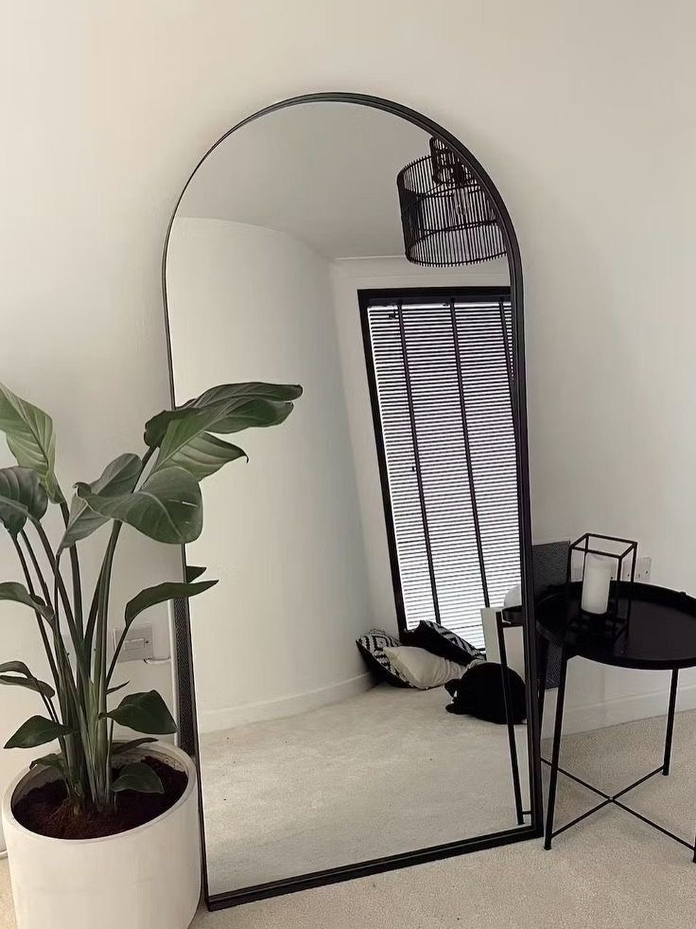 An arched black full-length mirror leaning against a white wall next to a potted plant.