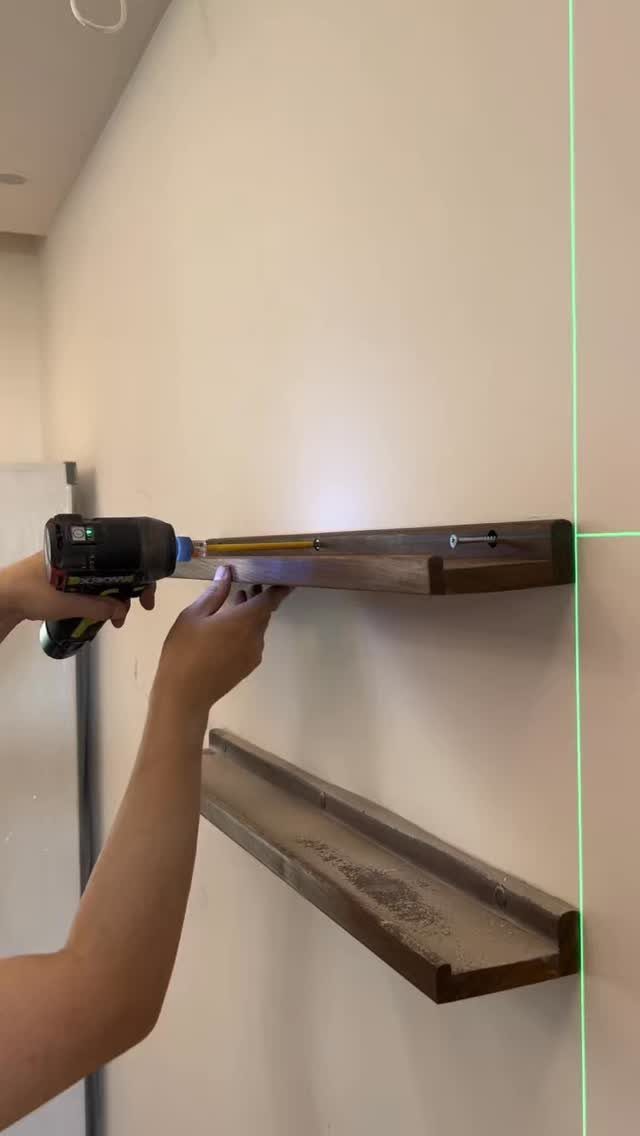 Hands installing a dark wooden floating shelf on a white wall using a power drill.