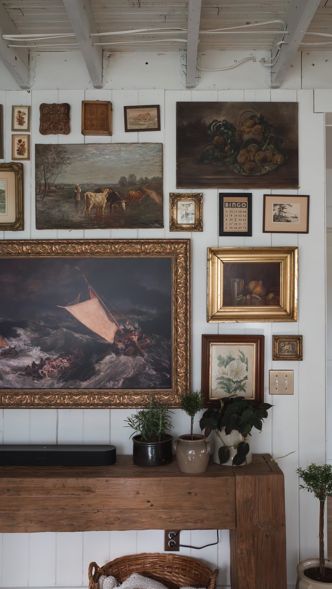 Assorted framed landscape and still life paintings arranged on a white paneled wall above a wooden table.