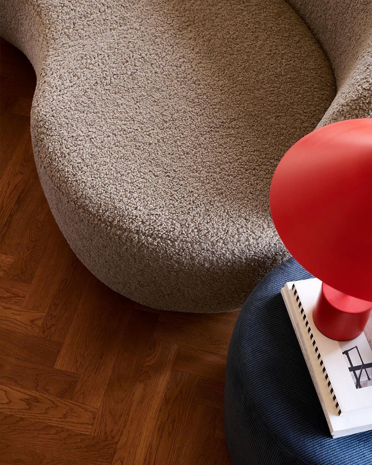 A curved beige bouclé chair on a wooden floor next to a red lamp.